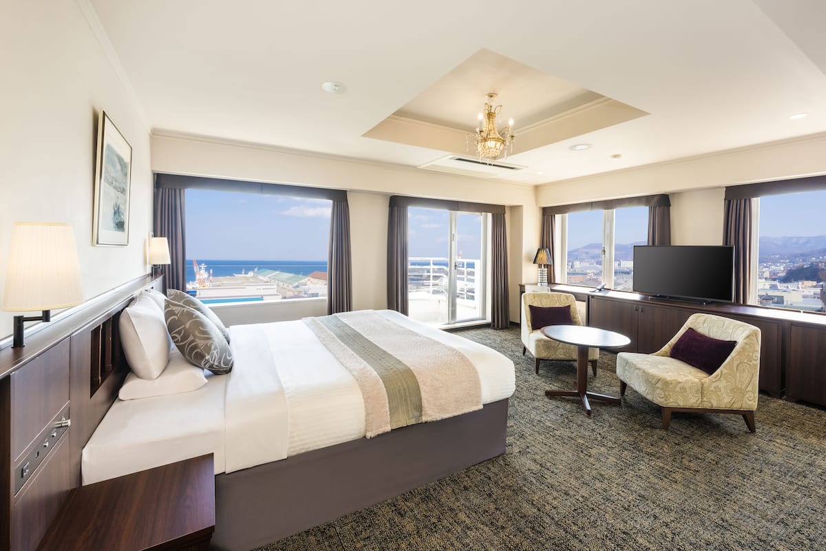 A hotel room with a large bed, a TV, a seating area, and a view of the city and ocean.