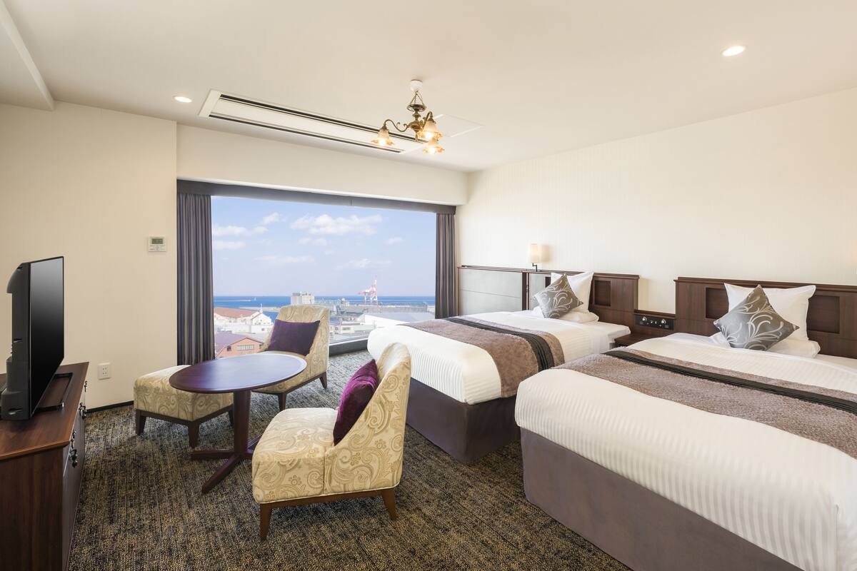 A hotel room with a large bed, a smaller bed, a desk with a TV, a chair, and a view of the city.