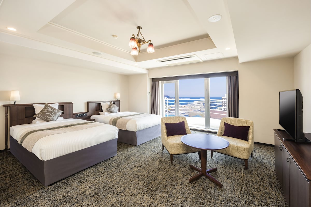 A hotel room with two beds, a TV, a small table, and a view of the ocean.