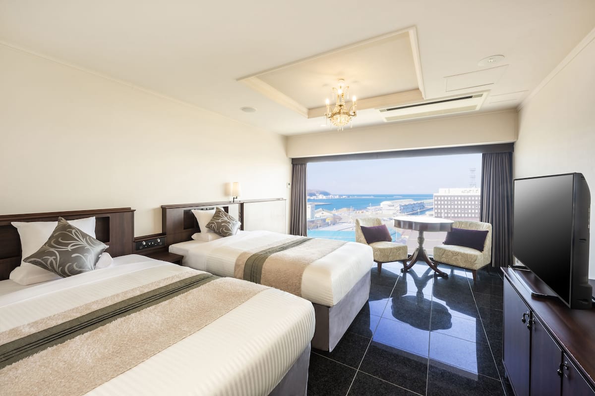 A hotel room with two beds, a TV, a balcony with a view of the city and ocean, and a chandelier.