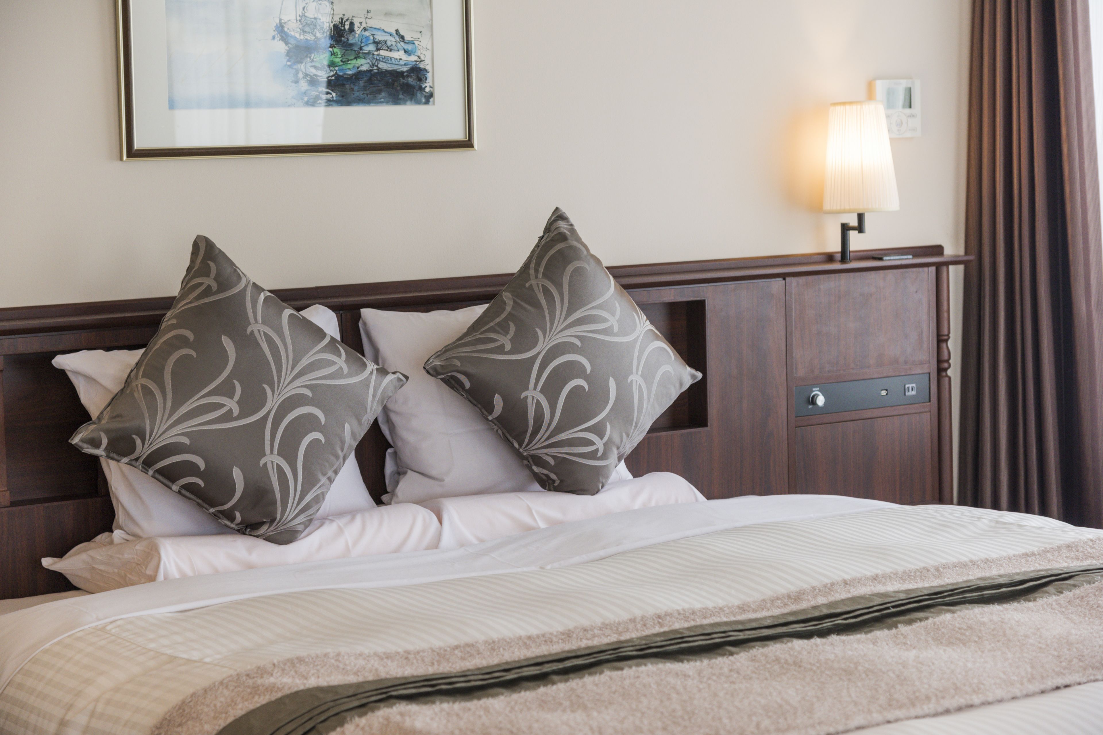 Een net opgemaakt bed met twee decoratieve kussens, een nachtkastje met een lamp en een lijst met een foto aan de muur.