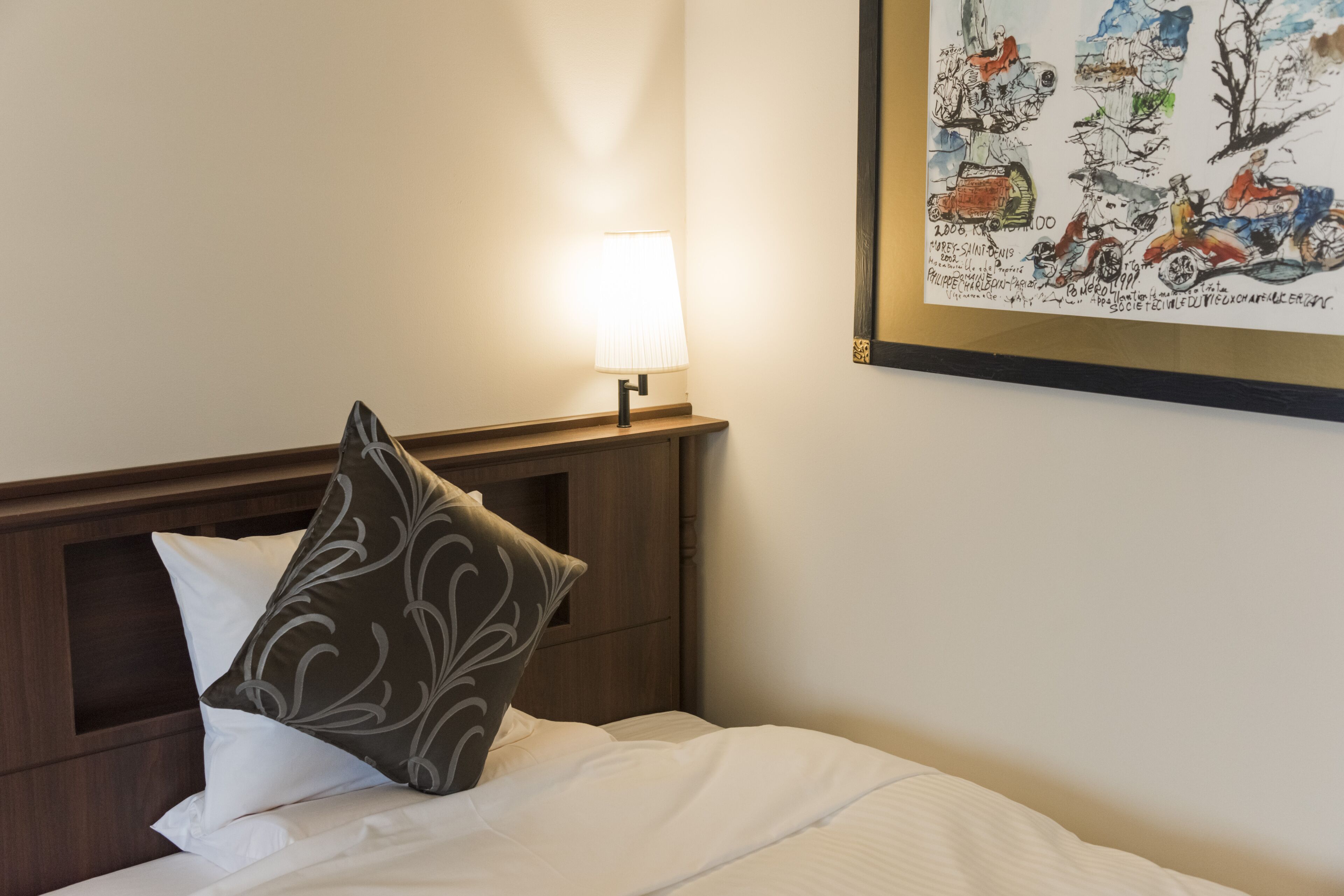 A neatly made bed with white linens and a brown pillow, a bedside lamp, and a framed artwork on the wall.