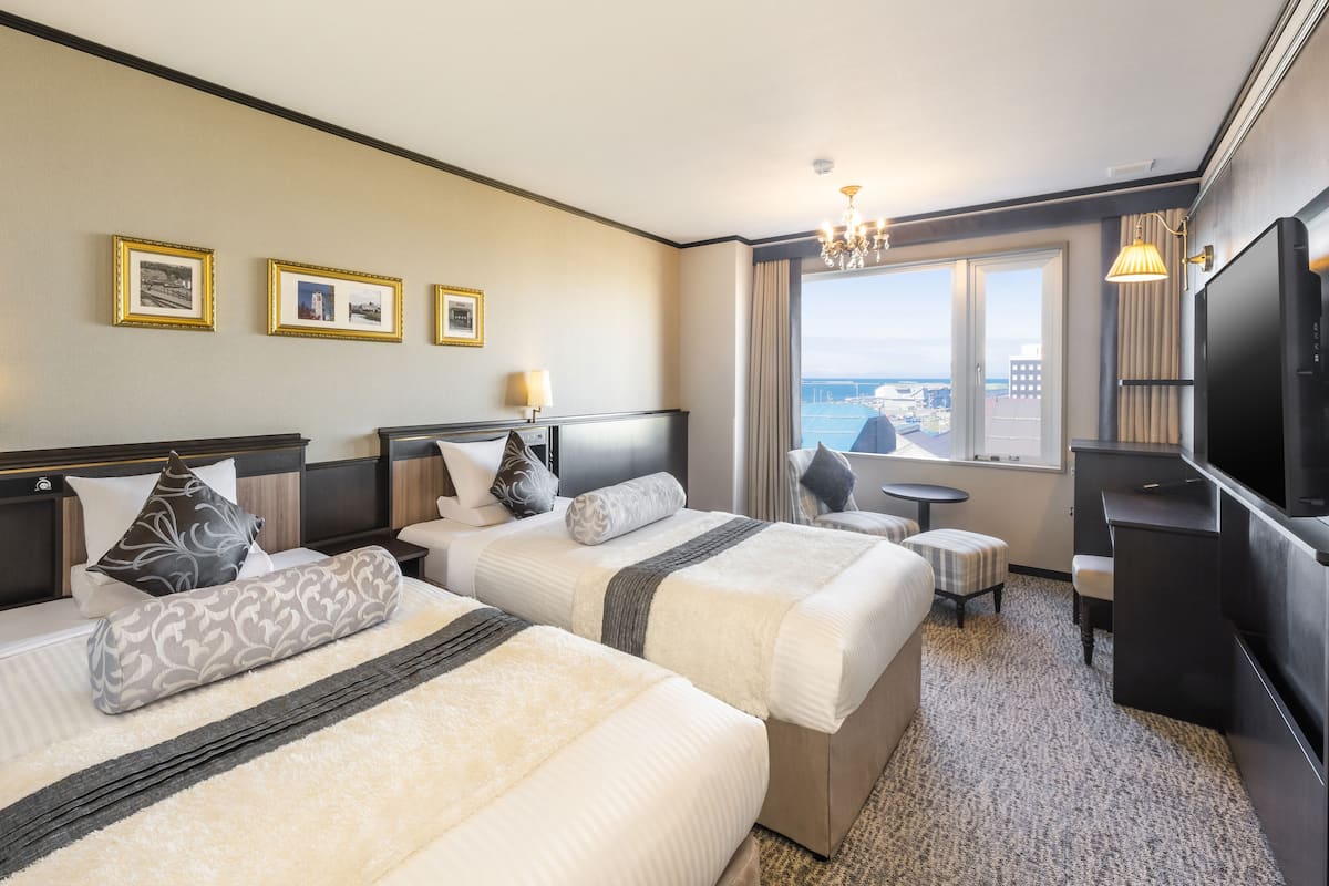 A hotel room with two beds, a desk, a TV, and a view of the city.