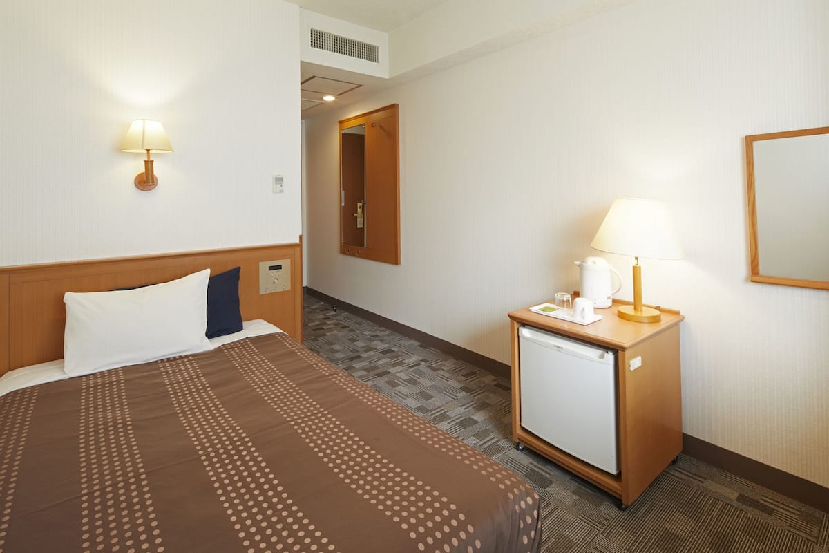 Une chambre d’hôtel avec un lit, une table de chevet avec une lampe, un petit réfrigérateur et un miroir au mur.