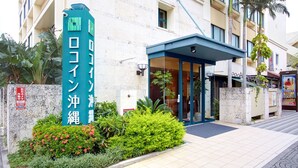 Entrada de hotel con un letrero verde, acceso cubierto y un pequeño jardín.
