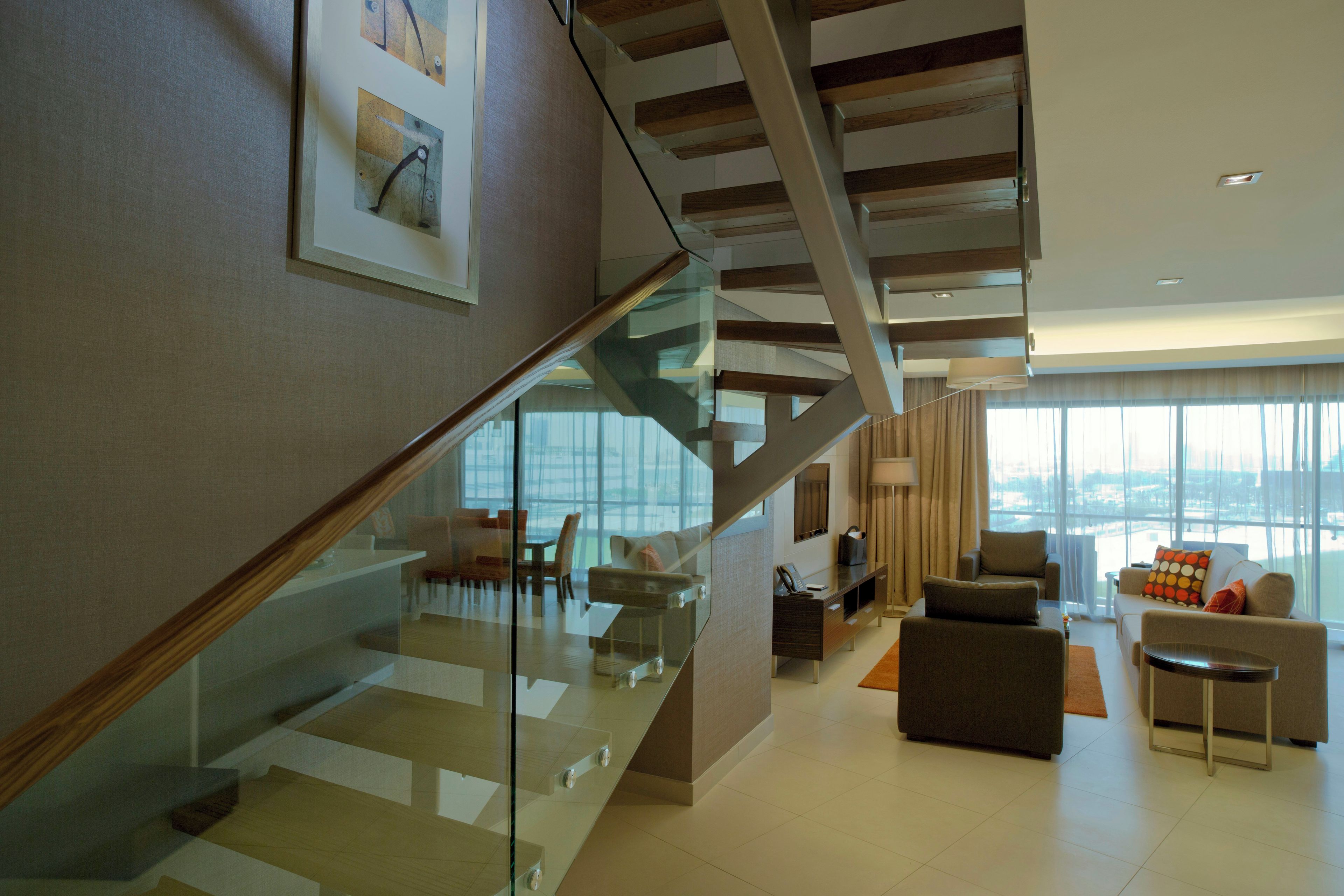Un interior moderno con una escalera de cristal, una zona de estar con sofás y vistas amplias a través de grandes ventanales.