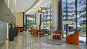 A modern lobby with a polished floor, orange seating, large windows, and indoor plants.