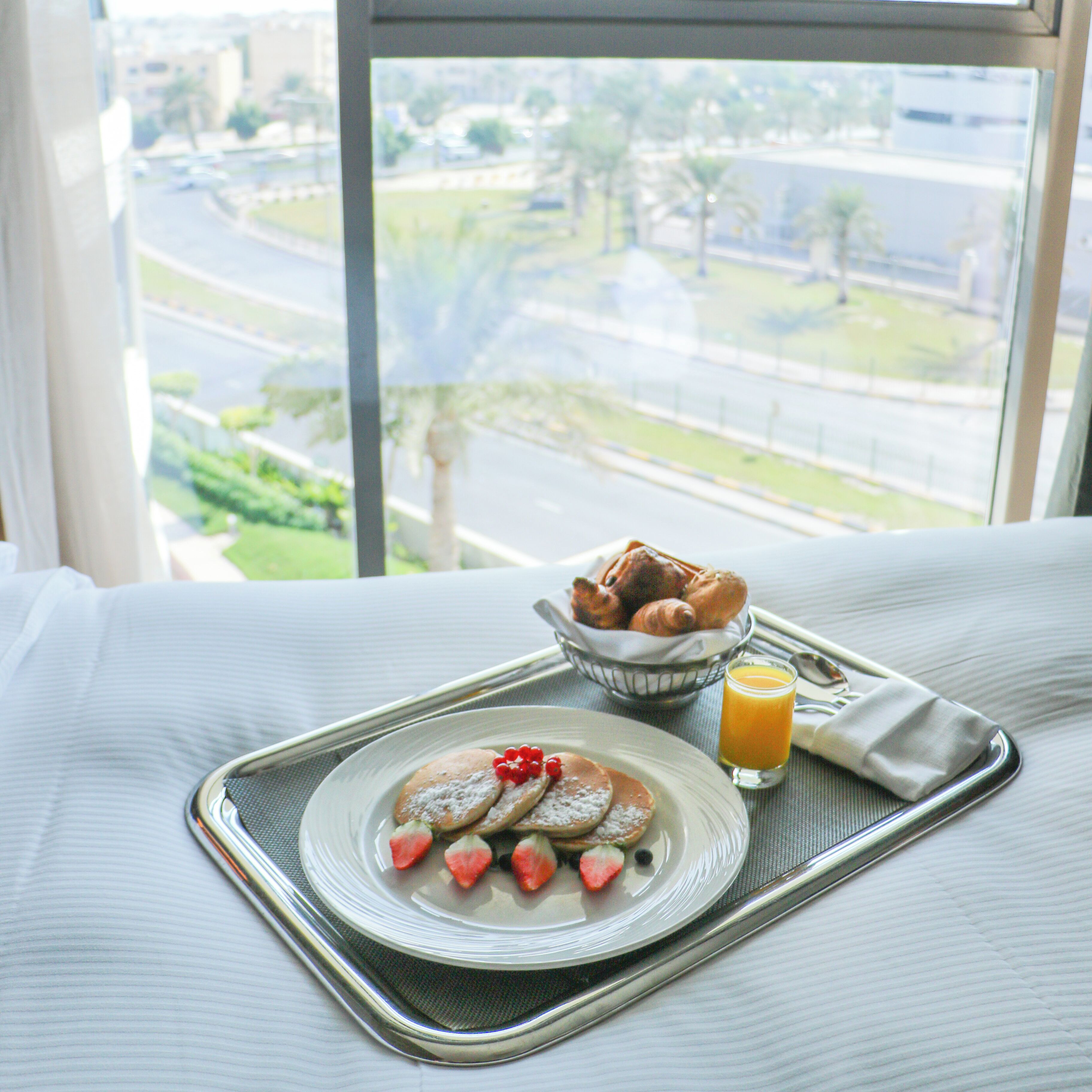 A breakfast tray with pancakes, strawberries, and a glass of orange juice on a bed with a window view of a cityscape.