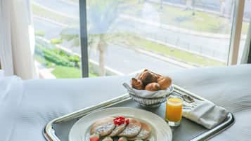 A breakfast tray with pancakes, strawberries, and a glass of orange juice on a bed with a window view of a cityscape.