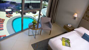A hotel room with a large window overlooking a swimming pool, a bed with a pillow, a bedside table with a lamp, and a chair.
