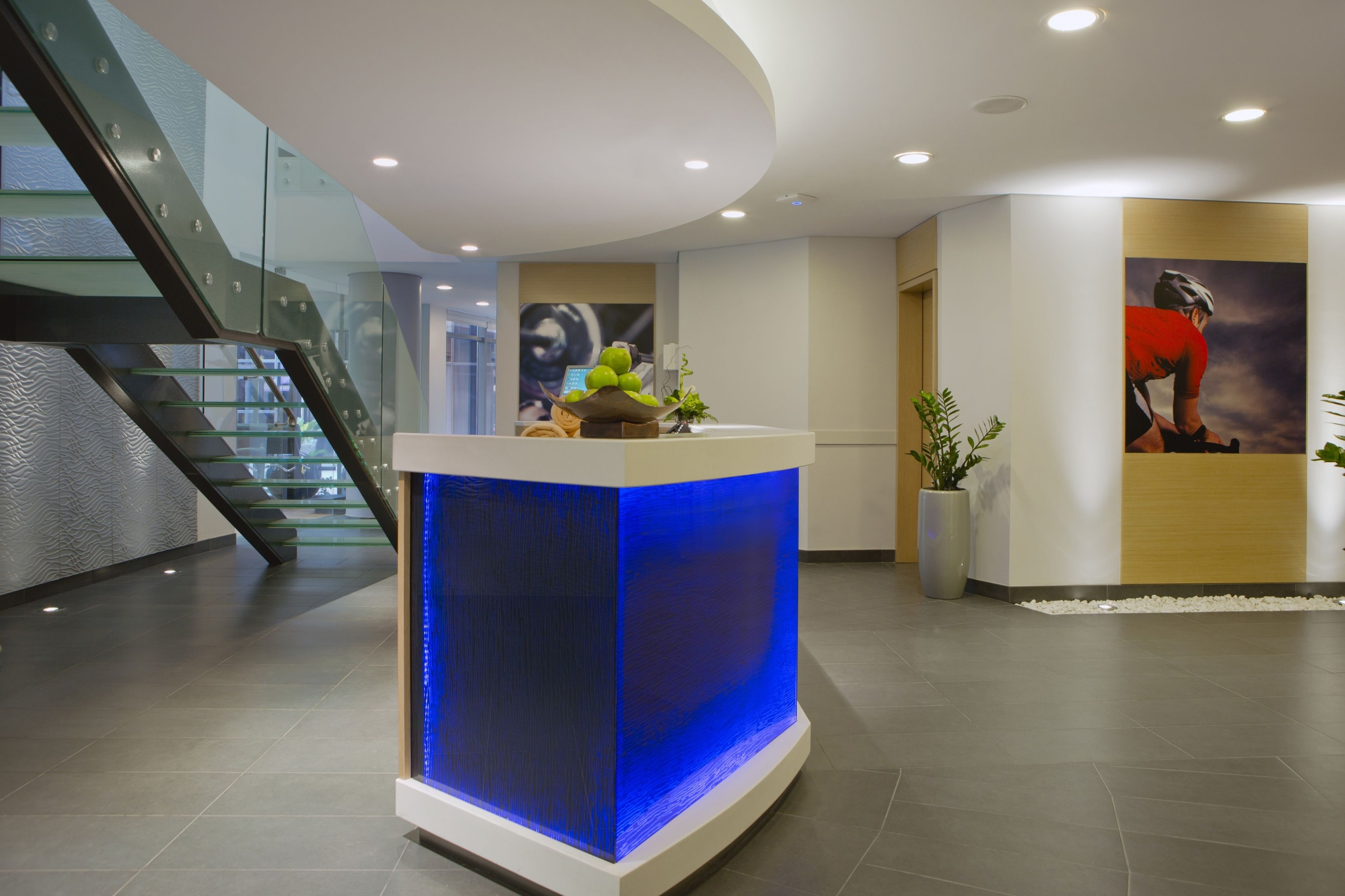 A reception area with a modern design, featuring a glass staircase, a reception desk with a blue illuminated panel, and a poster of a cyclist.