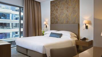 Ein Hotelzimmer mit einem großen Bett, einem Schreibtisch mit Stuhl, einem Nachttisch mit Lampe und Blick auf ein Gebäude durch ein Fenster.