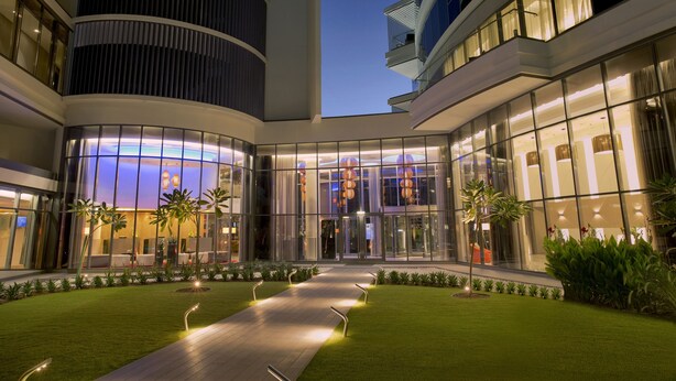 A modern hotel with a glass facade, a central courtyard with a pathway, and a well-maintained lawn.