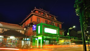 Un edificio tradicional con un letrero verde llamativo, un área cubierta con techo y una calle con estelas de luz que indican movimiento.