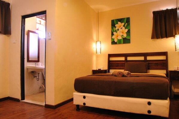 A hotel room with a bed, a bedside table, a nightstand, a wall-mounted light fixture, and a painting of flowers. - Bemo Corner Guest House (Kuta)