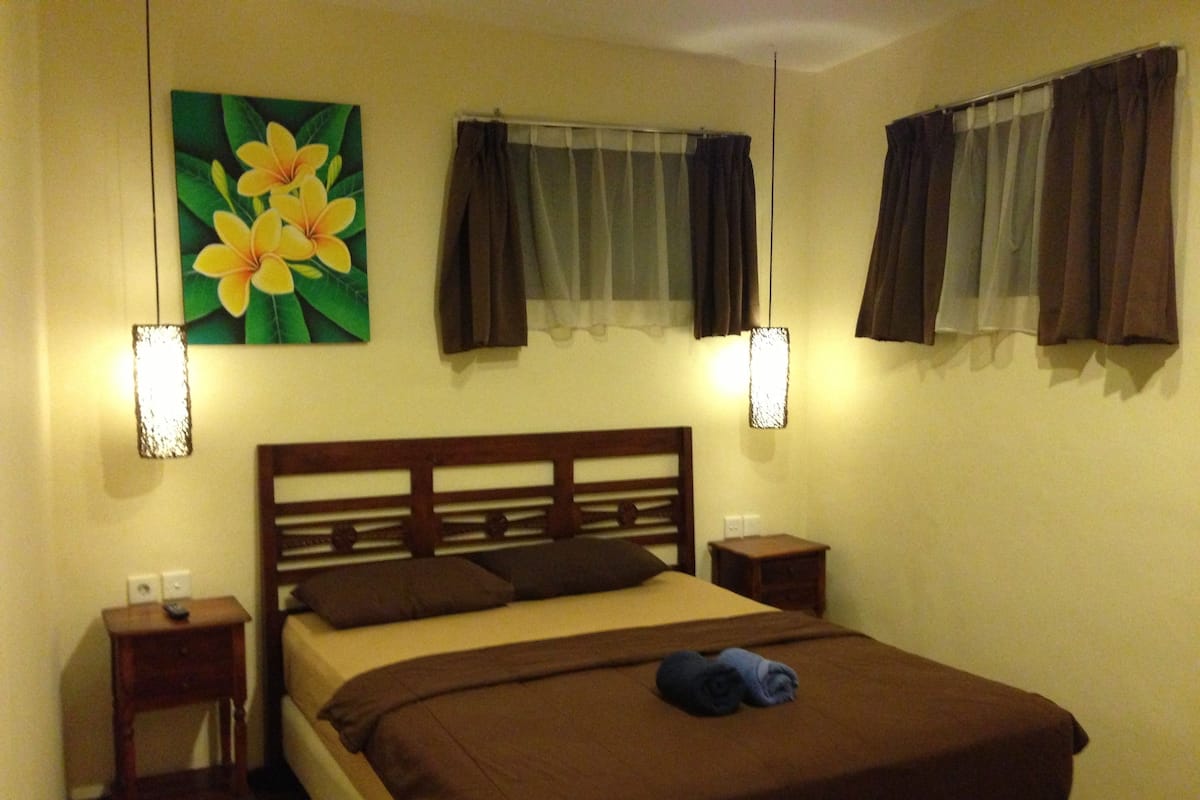 A bedroom with a wooden headboard, a bed with brown bedding, two bedside tables, a nightstand, a wall-mounted lamp, and a floral painting.