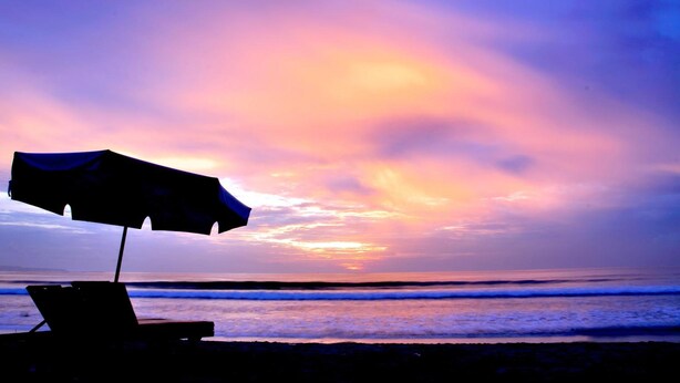 Escena de playa con atardecer, una sombrilla y una silla de descanso.