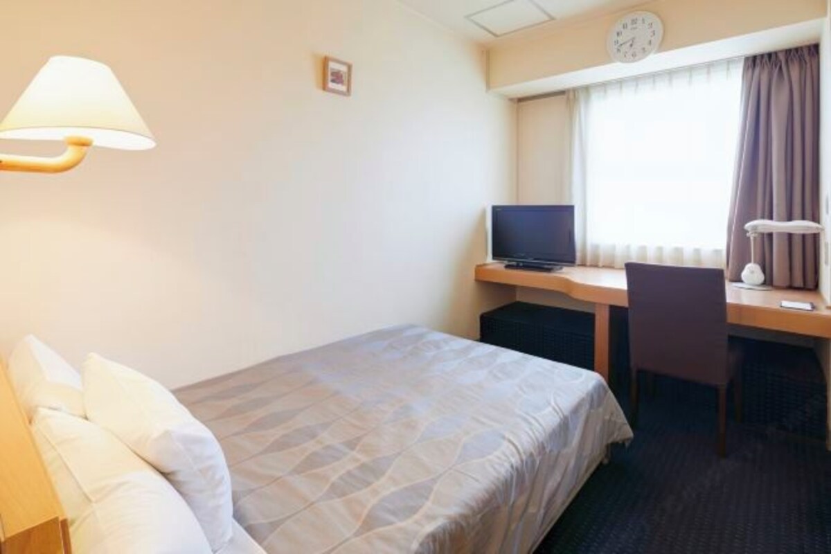 Ein Hotelzimmer mit einem Bett, einem Schreibtisch mit Computer, einem Stuhl, einer Lampe und einer Wanduhr.