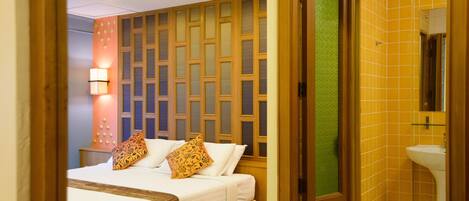 A hotel room with a bed, a nightstand, a wall-mounted lamp, and a bathroom with a sink and mirror.