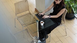 Eine Frau sitzt an einem Glastisch mit einem Laptop, umgeben von Korbstühlen und einem Blumentopf.