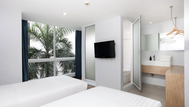 Ein modernes Hotelzimmer mit zwei Betten, einem Fernseher, einem Badezimmer inklusive Spiegel und Waschbecken sowie einem großen Fenster mit Blick auf Palmen.