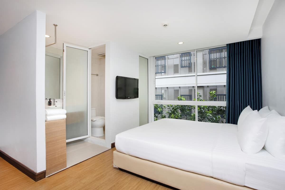 A modern hotel room with a bed, a bathroom, a TV, and a balcony with a view of greenery.