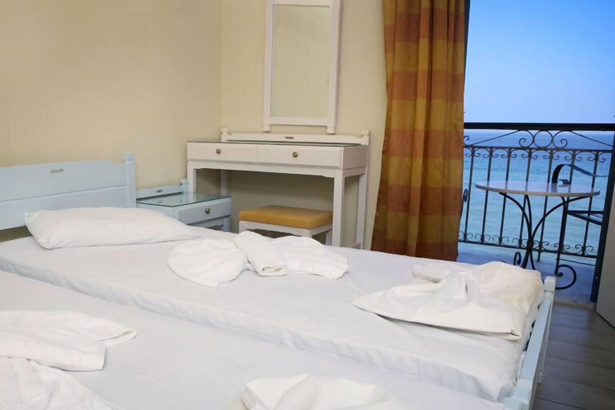 A hotel room with two beds, a small table with a chair, a bench, a mirror, and a view of the sea through a balcony.
