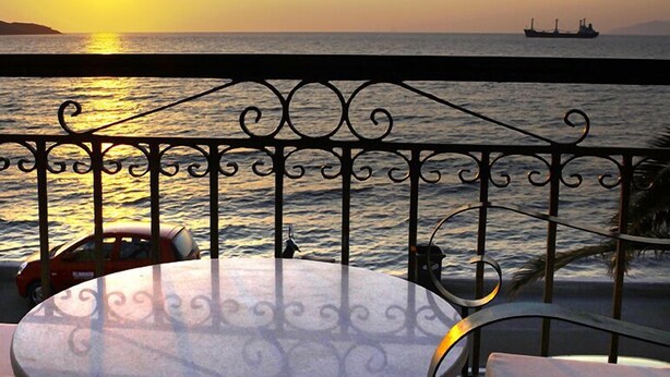 Un balcón con vista al mar al atardecer, que incluye una mesa redonda y sillas, y una barandilla metálica decorativa.