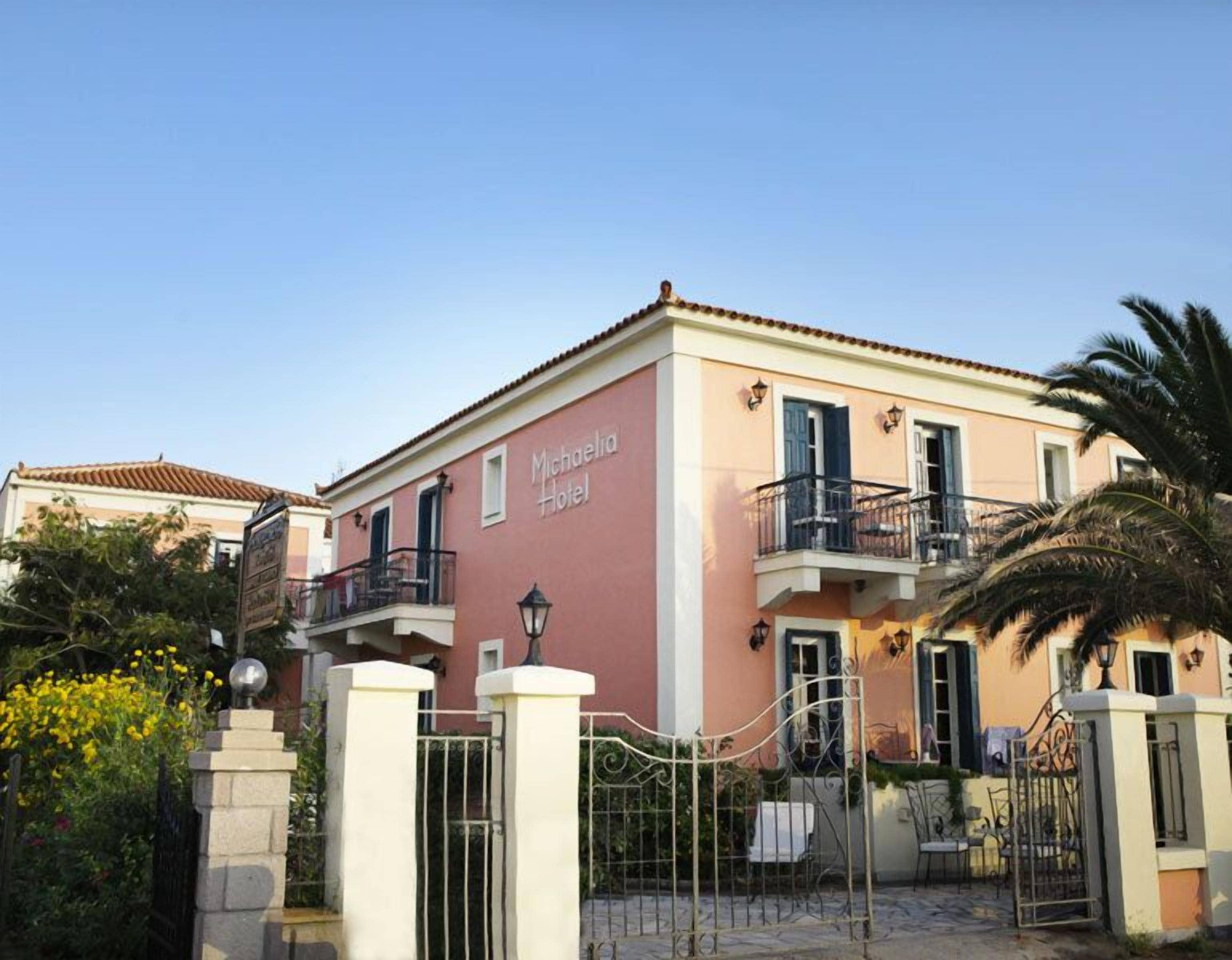 Un edificio de dos pisos con fachada rosa, llamado 'Michaelia Hotel'. Cuenta con un balcón con barandilla negra, una entrada con portón y una pequeña zona de estar al aire libre.