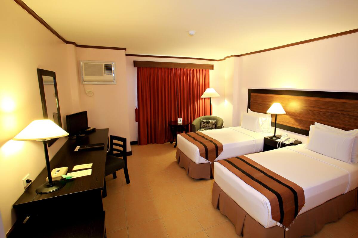 A hotel room with two beds, a desk with a computer, a chair, a lamp, and a window with curtains.