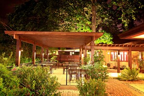 An outdoor patio area with wooden tables and chairs, surrounded by lush greenery and potted plants.