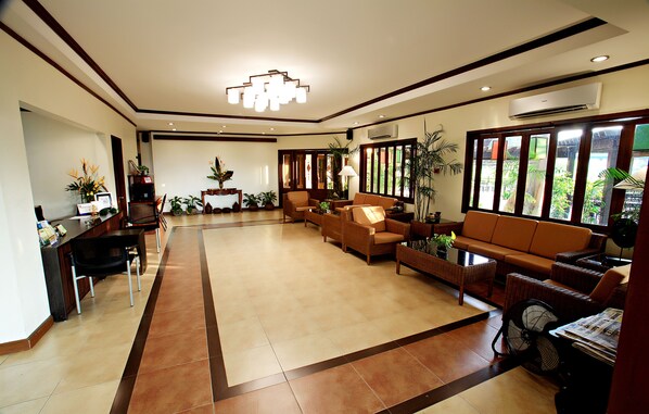 A spacious living area with a sofa set, a coffee table, and a dining table with chairs. There are potted plants and a fan on the right side.
