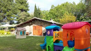 Une aire de jeux pour enfants dans un jardin, avec une maison en pierre en arrière-plan.