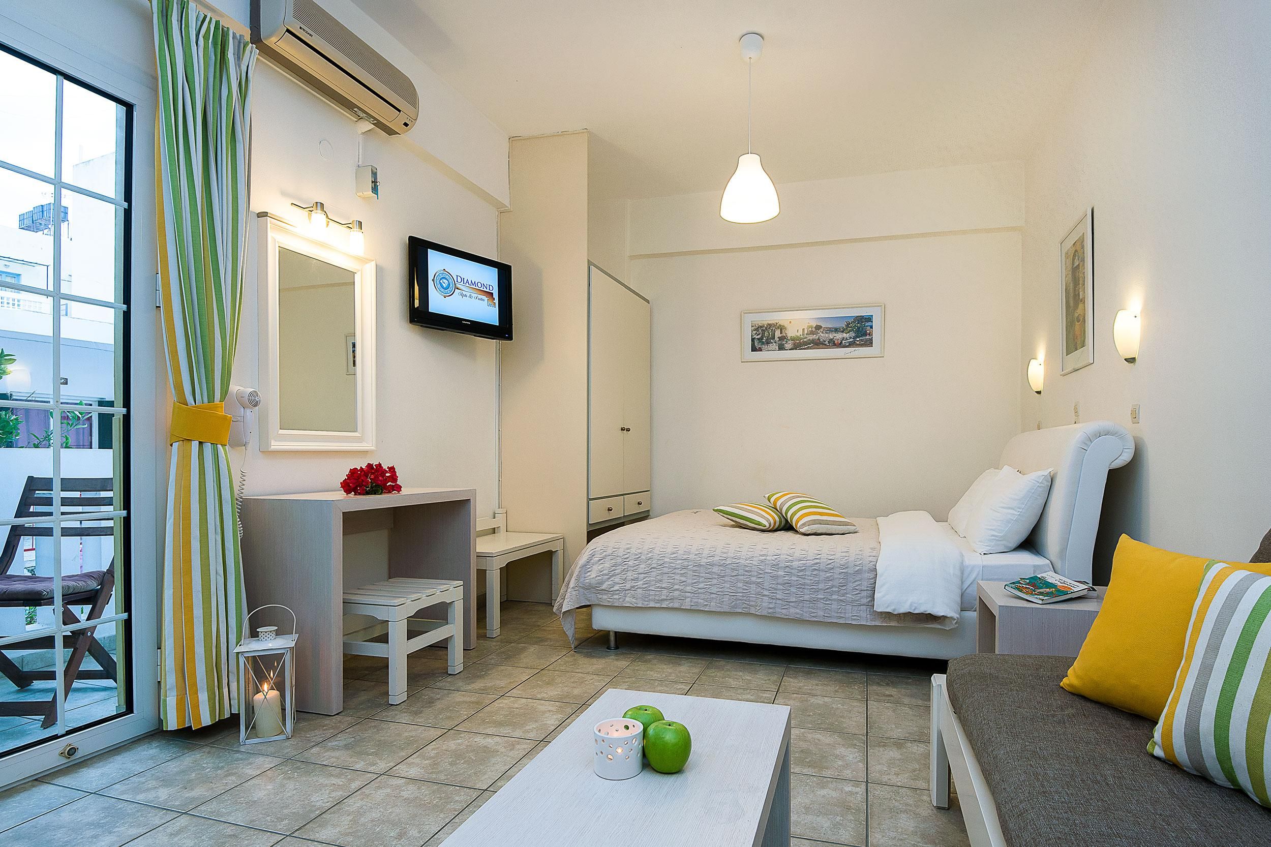 Una habitación de hotel con una cama, un escritorio, una silla, una mesa con manzanas y una vela, y un televisor en la pared.