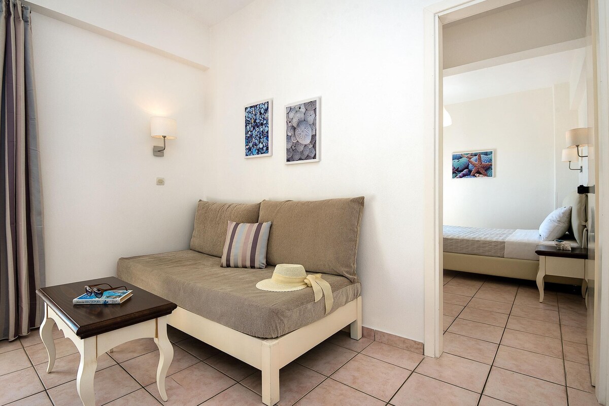 A cozy room with a sofa, a small table, and a bed. There are wall-mounted lights and framed pictures on the wall.