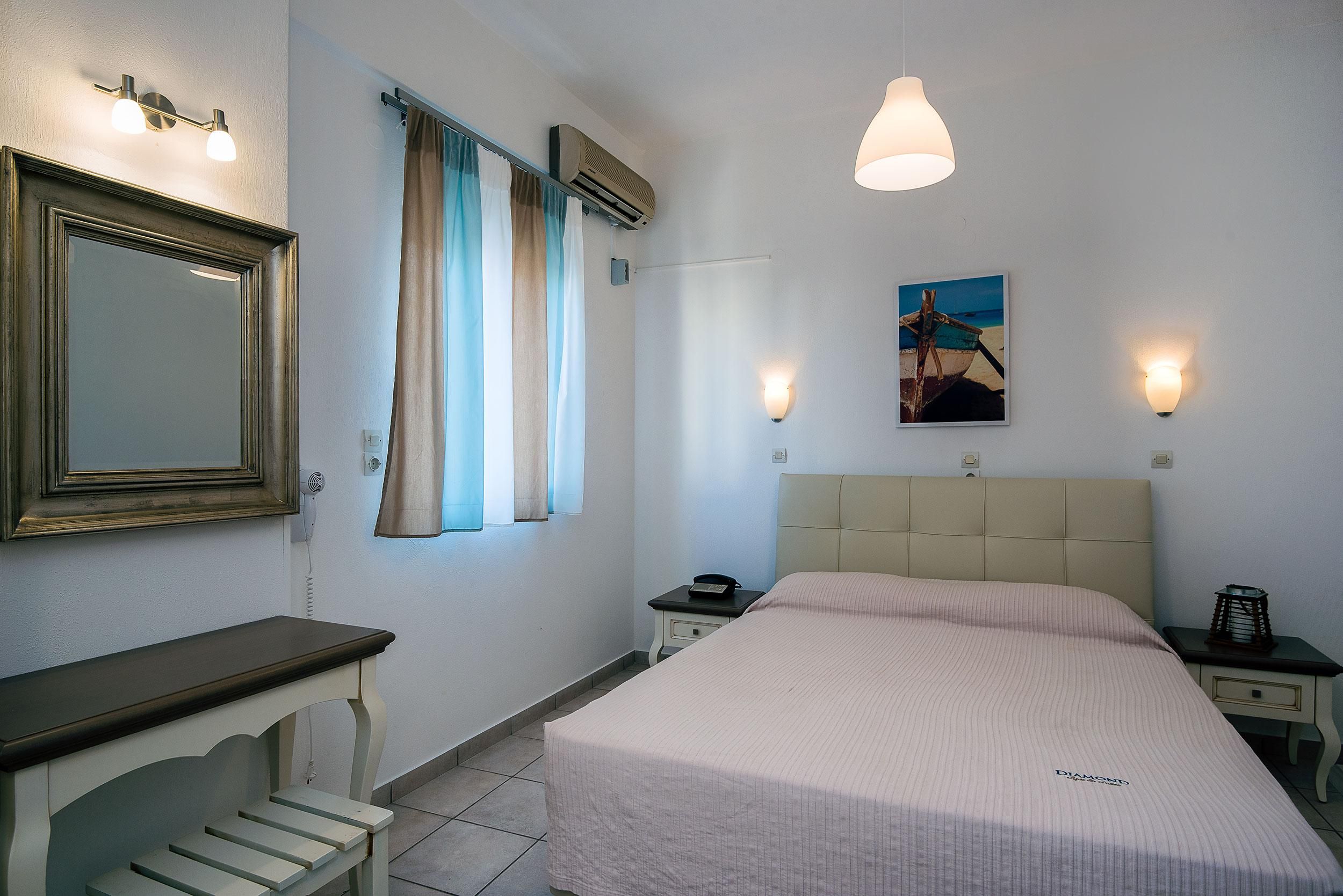 Una habitación de hotel con una cama, una mesita de noche, un espejo, una ventana con cortinas, un aire acondicionado, una lámpara de pared y un cuadro colgado.