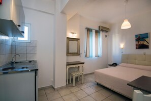 A compact hotel room with a kitchenette, a bed, a small table, a chair, a mirror, and a window with curtains. - Diamond Apts and Suites (Hersonissos)