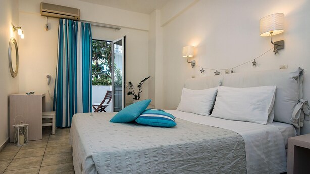 A bedroom with a bed, a nightstand, a lamp, a mirror, and a window with curtains. - Diamond Apts and Suites (Hersonissos)