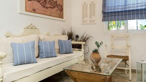A white sofa with blue pillows, a glass coffee table, a map on the wall, a window with a blue blind, and a white chair. - Diamond Apts and Suites (Hersonissos)