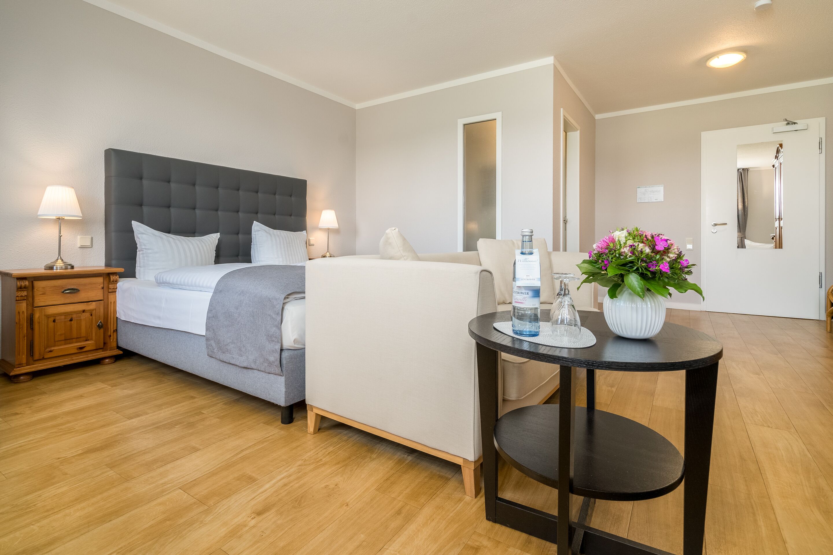 Een hotelkamer met een bed, nachtkastje, lamp, stoel en een kleintje tafeltje met een bloemenvaas.