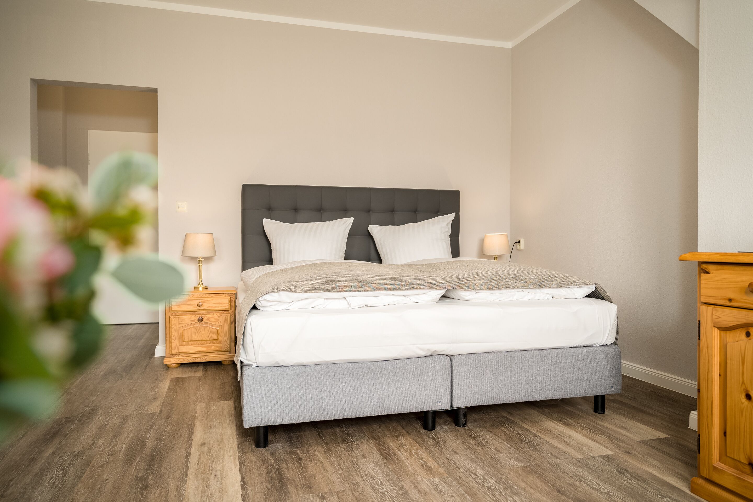 Une chambre à coucher avec un lit recouvert d’un tissu gris, deux tables de chevet avec des lampes et une table de nuit en bois.
