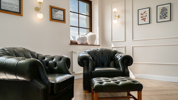 Een kamer met twee zwarte leren fauteuils en een bijpassend poefje, een raam met decoratieve vazen, en wandlampen.