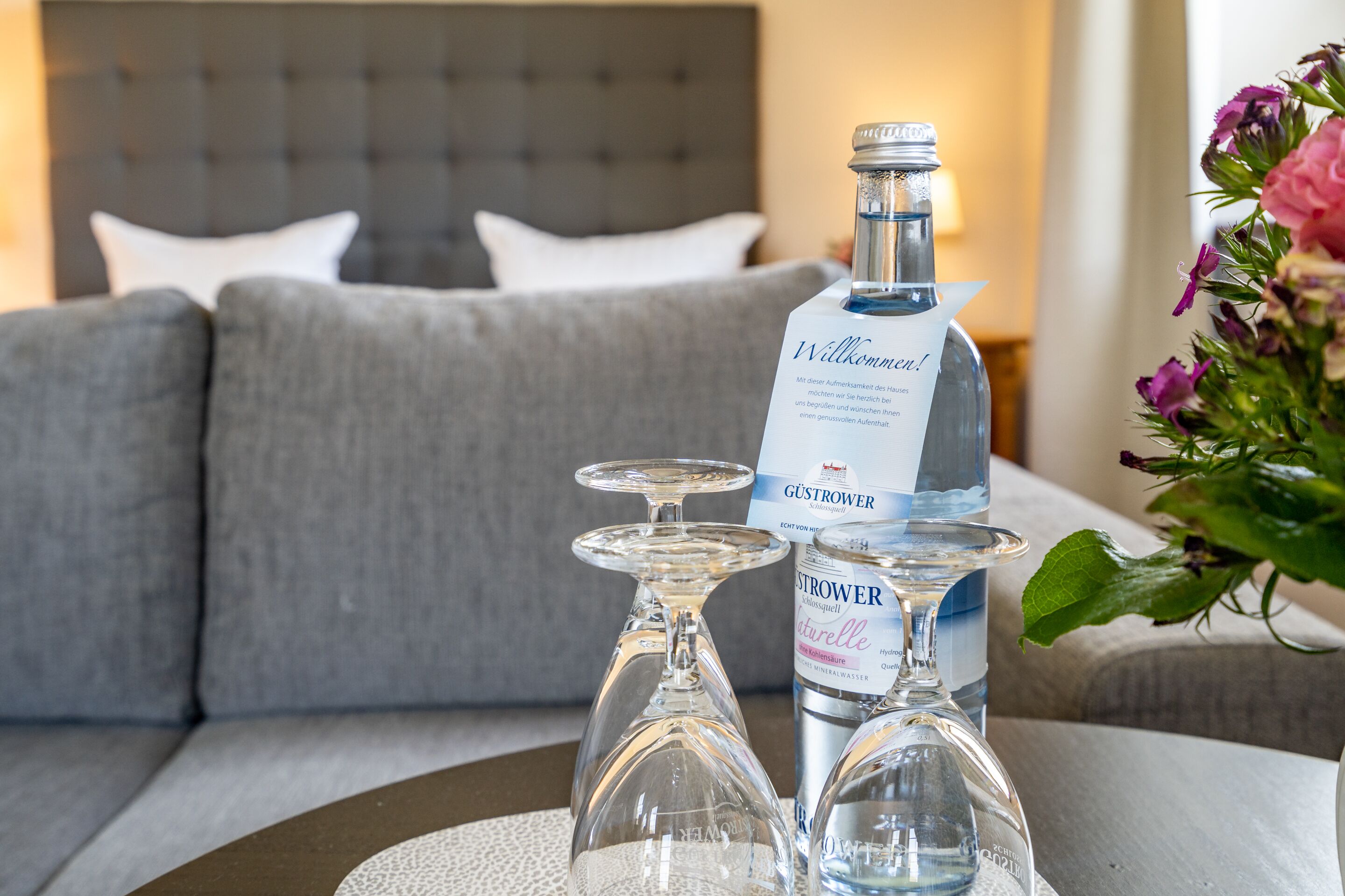 Una botella de agua, un vaso y un juego de vasos sobre una mesa en una habitación de hotel con un sofá gris y un ramo de flores.
