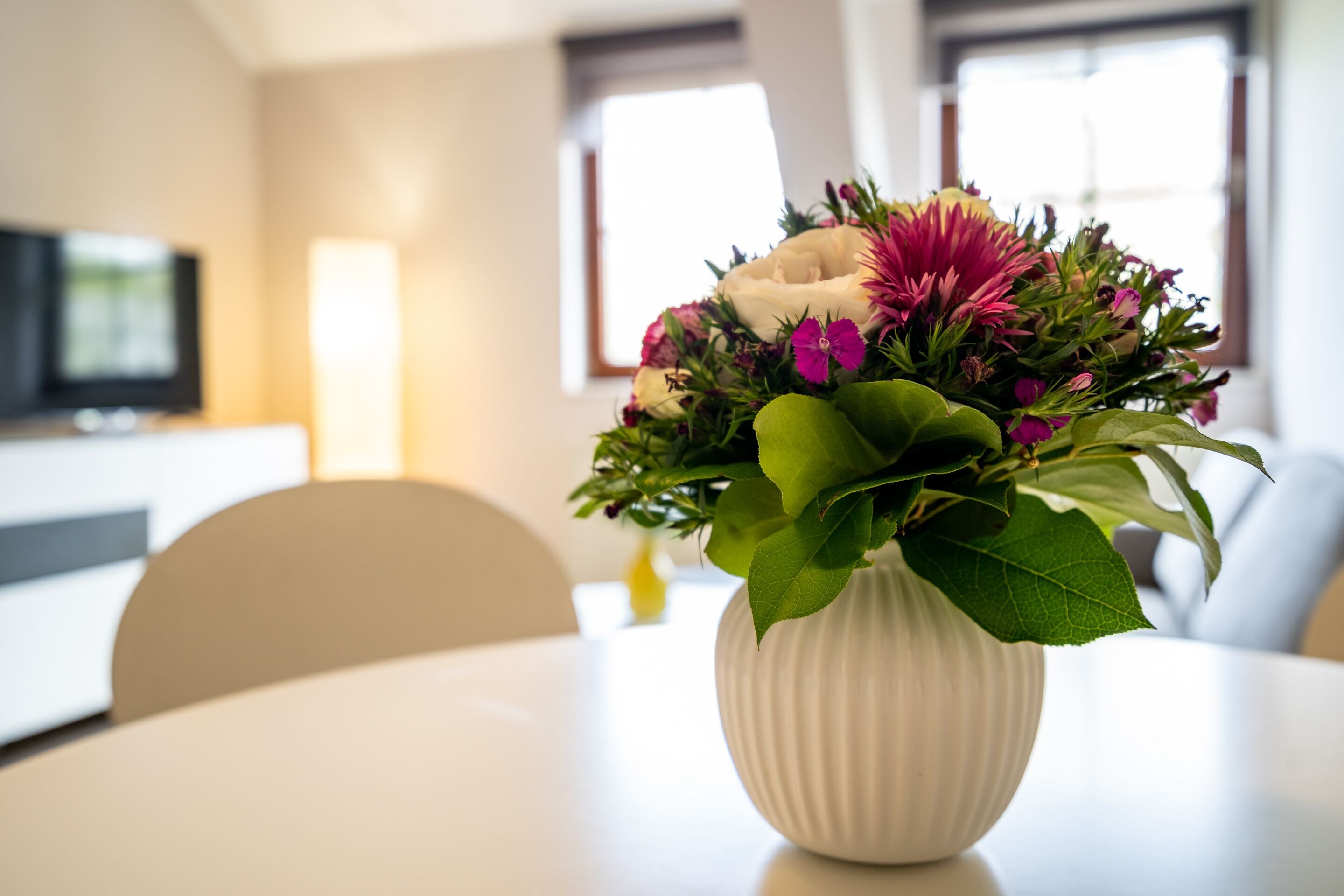 A white vase with a floral arrangement on a table in a room with a television in the background.
