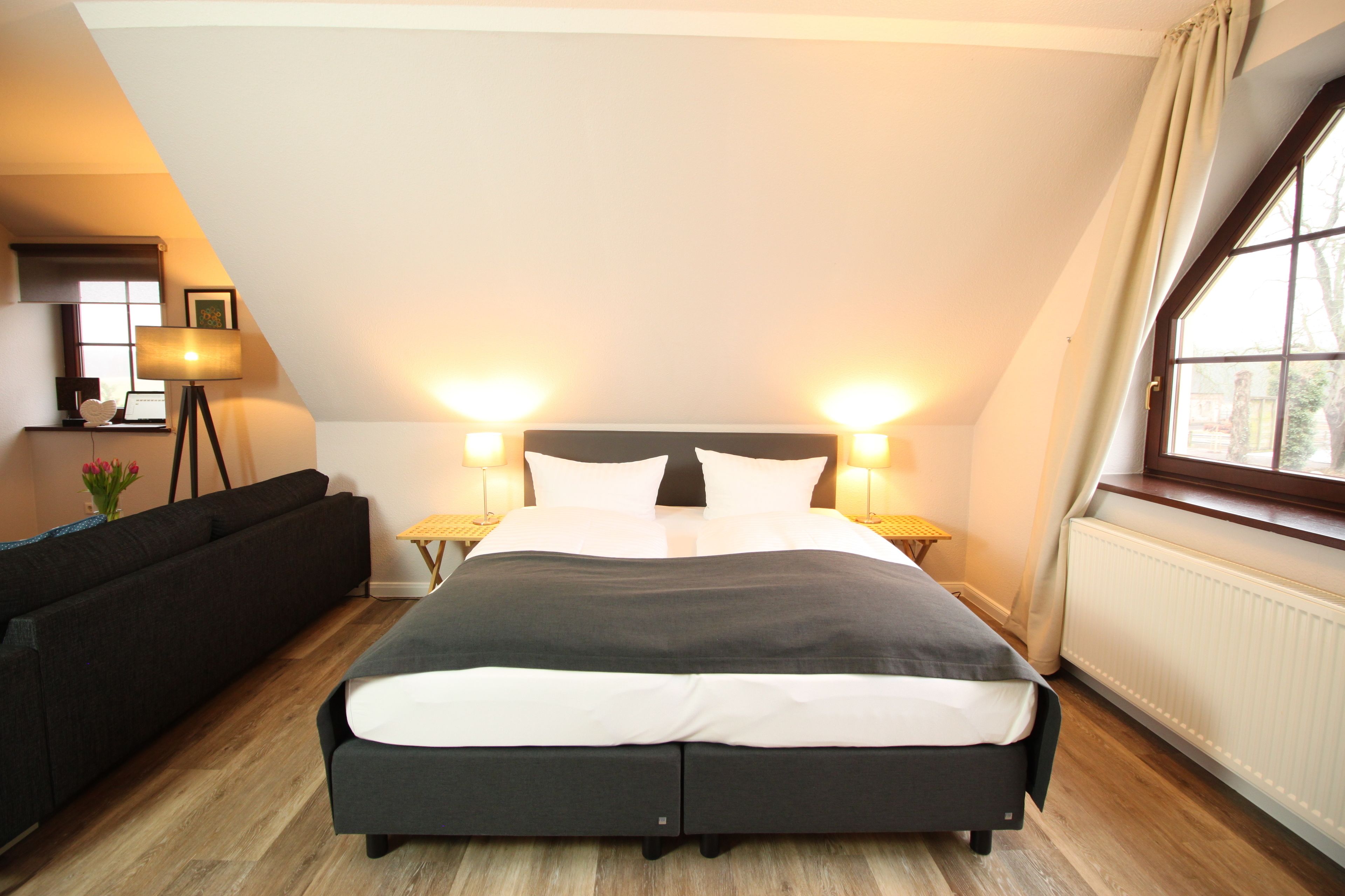 A bedroom with a large bed, two bedside lamps, a window with curtains, a radiator, and a sofa in the corner.