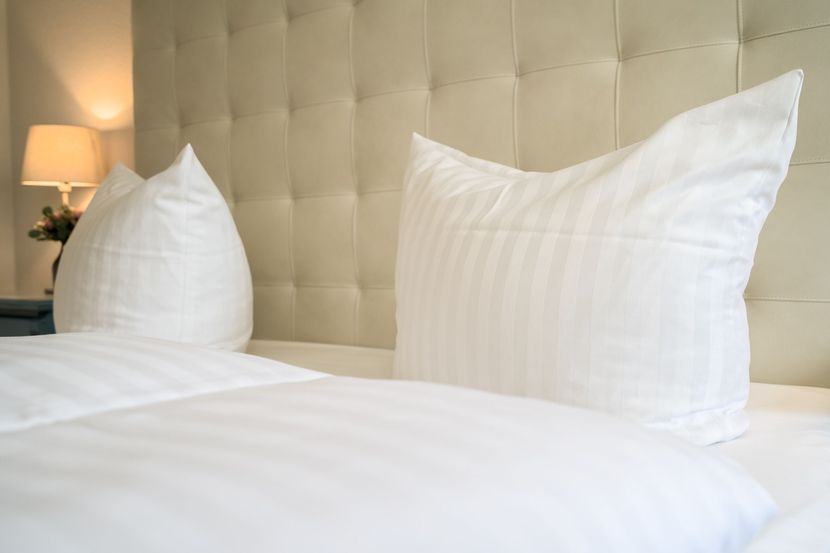 Een net opgemaakt bed met witte lakens en kussens, een gestoffeerd hoofdbord en een bedlampje.