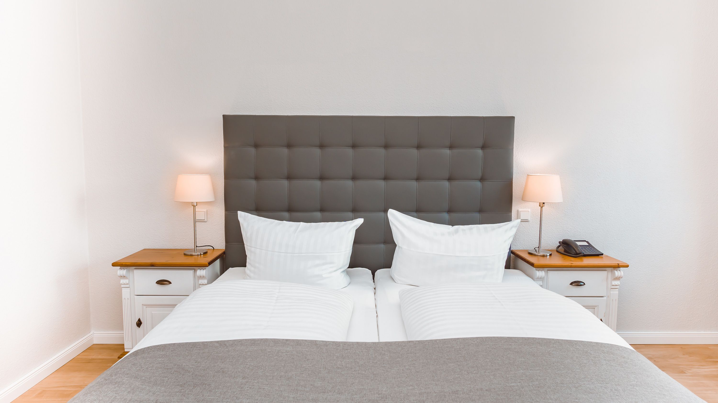Een opgemaakt bed met een grijze hoofdbord, twee nachtkastjes met lampen, en een houten bijzettafeltje.