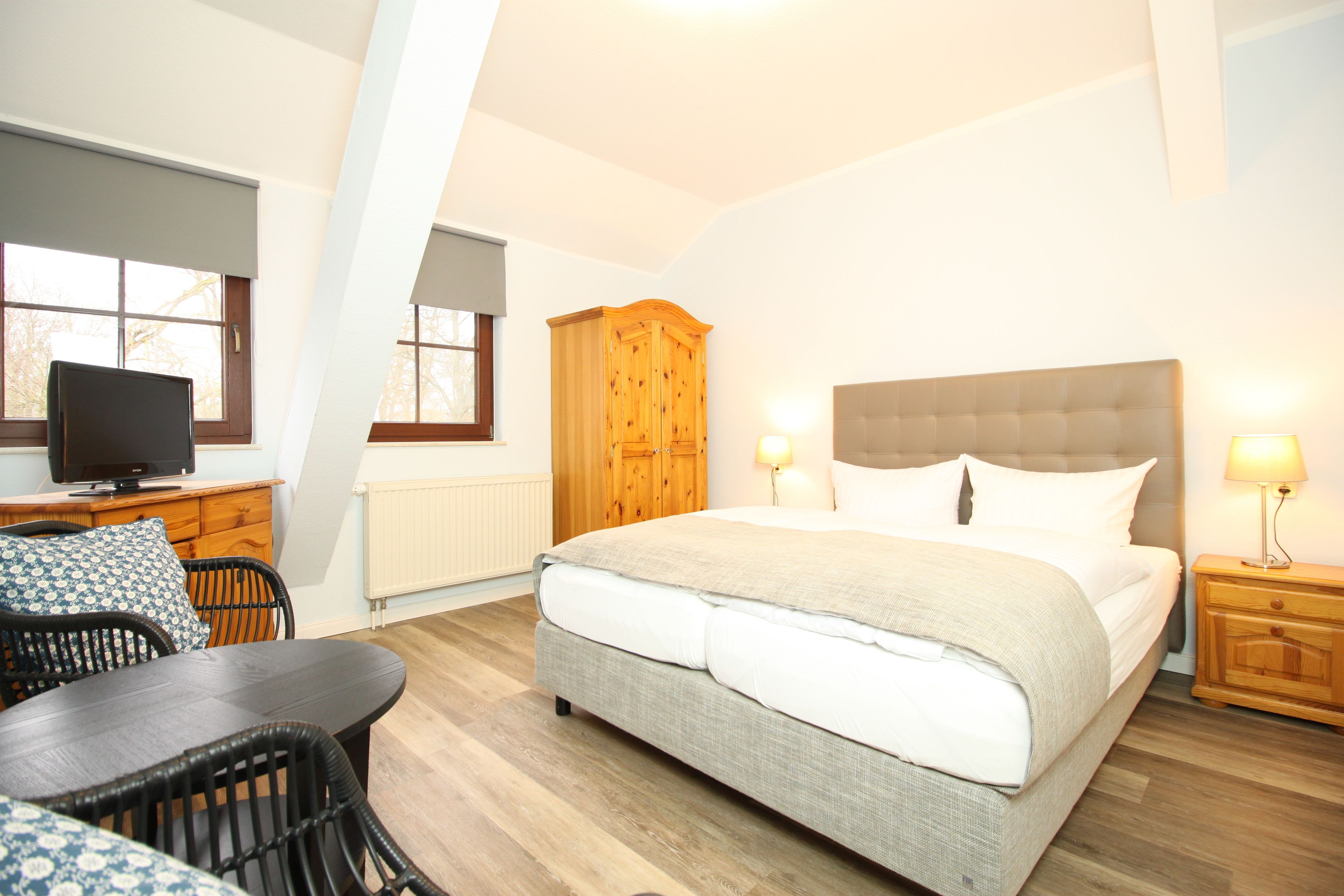 A bedroom with a bed, a wooden wardrobe, a television, a table with chairs, and a window.