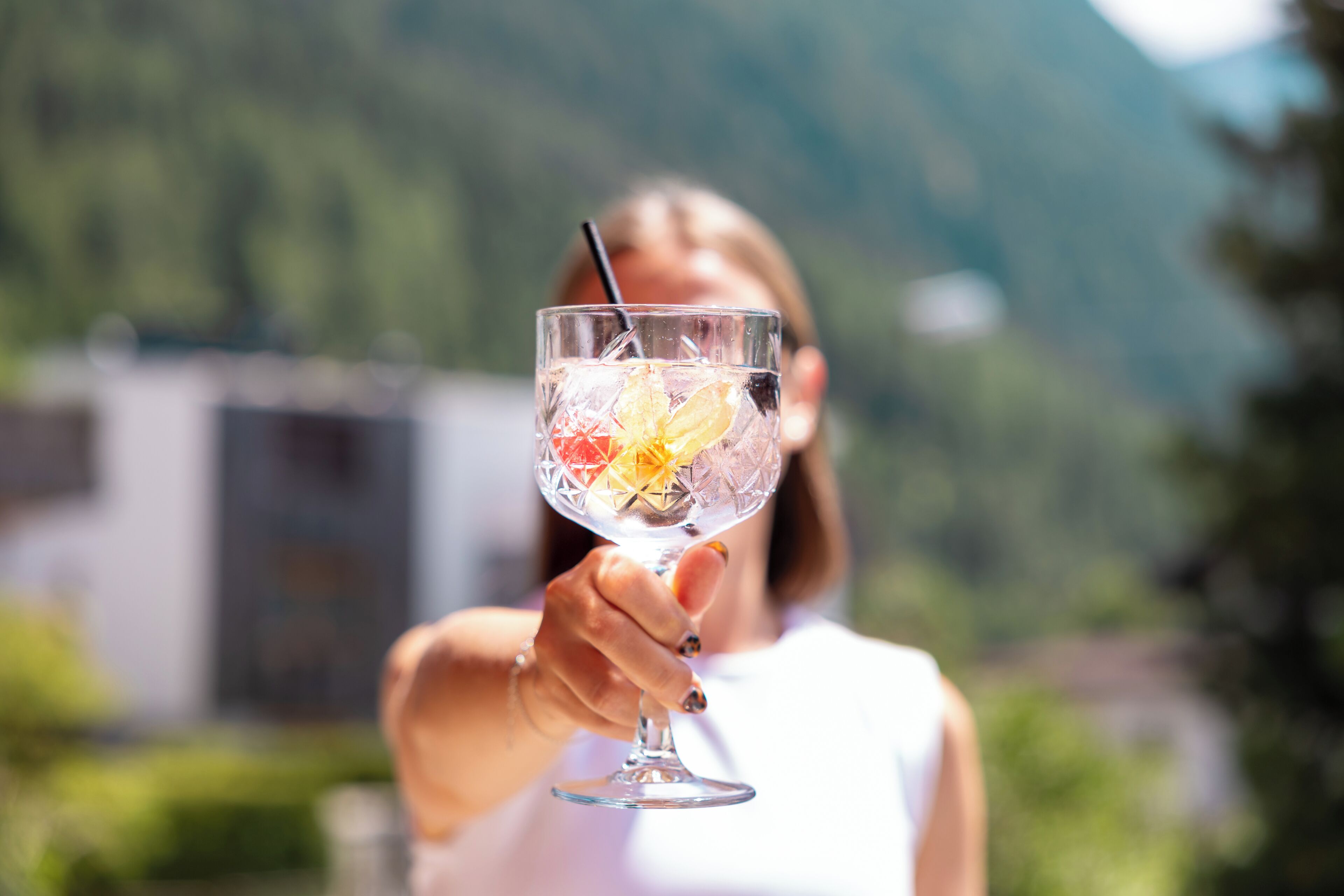 Una persona sosteniendo un vaso transparente con pajita, que contiene una bebida y rodajas de fruta, al aire libre con una montaña y un edificio de fondo.