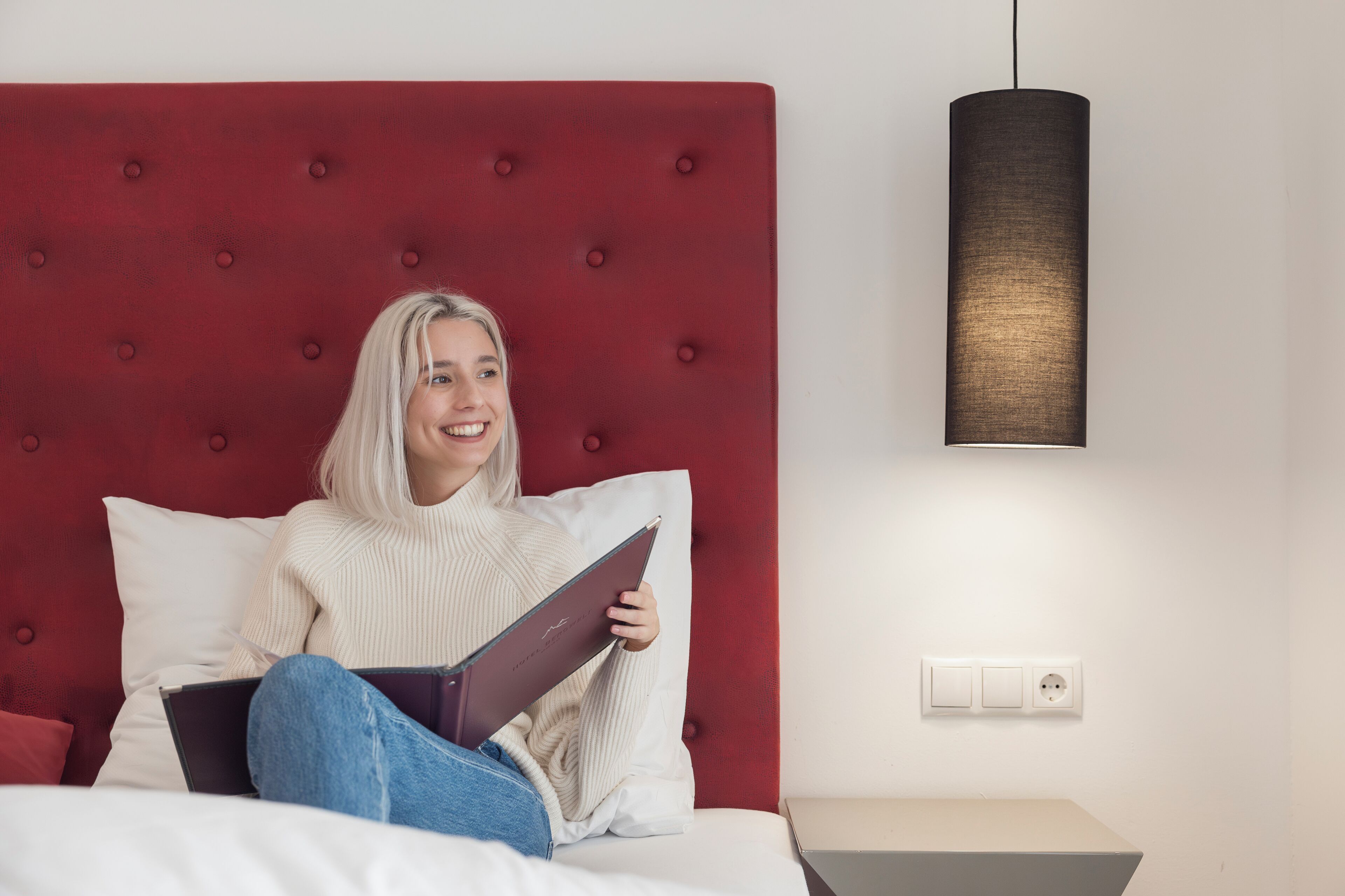 Una persona sentada en una cama con cabecera roja, sosteniendo un libro. Hay una mesita de noche con una lámpara y un enchufe en la pared.
